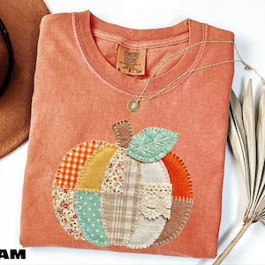 May include: A terracotta-colored t-shirt featuring a large, central pumpkin applique made from various fabric patches including gingham, floral, plaid, and lace patterns. The pumpkin has a light green fabric leaf and visible blanket stitching.