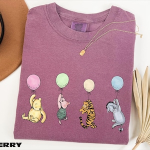 A purple t-shirt with illustrations of four cartoon characters from the Winnie the Pooh series, including Tigger, Tigger's friend, Tigger, and Eeyore, each holding a balloon. The t-shirt is displayed on a white background, along with a hat, a necklace, and some fans.
