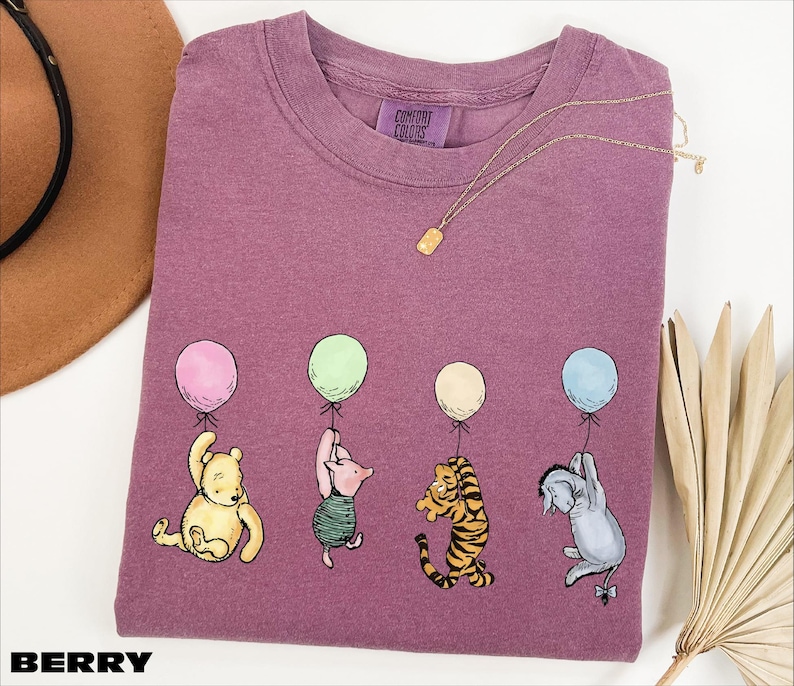 A purple t-shirt with illustrations of four cartoon characters from the Winnie the Pooh series, including Tigger, Tigger's friend, Tigger, and Eeyore, each holding a balloon. The t-shirt is displayed on a white background, along with a hat, a necklace, and some fans.