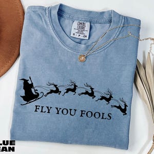 May include: A blue jean colored t-shirt with a black graphic of a wizard in a sleigh pulled by reindeer. The text "FLY YOU FOOLS" is below the graphic. A gold necklace with a pendant is on the shirt.