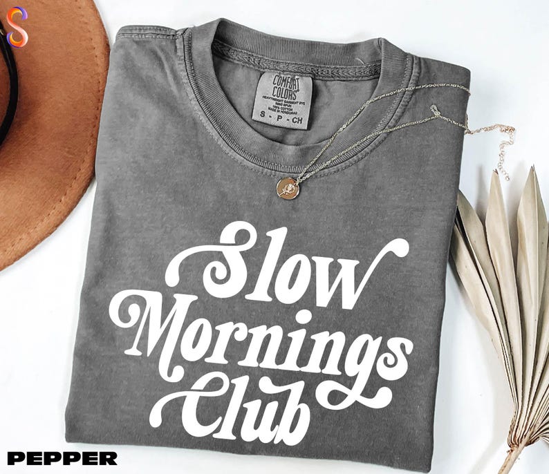 a gray t-shirt with the text "Slow Mornings Club" printed on it, along with a necklace and a hat.