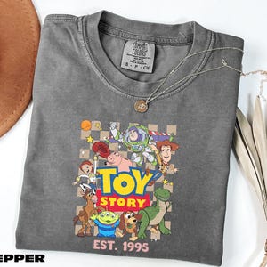A gray t-shirt with a colorful graphic of characters from the Toy Story movie, including Woody, Buzz Lightyear, and other beloved characters, along with the text "Toy Story" and "Est. 1995".