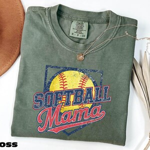 a green t-shirt with a graphic design featuring a baseball and the text "Softball Mama" printed on it. The t-shirt is folded and placed on a white surface, with a brown hat and some dried leaves nearby.