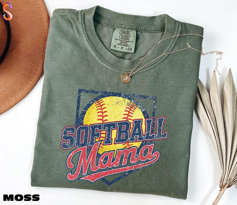 a green t-shirt with a graphic design featuring a baseball and the text "Softball Mama" printed on it. The t-shirt is folded and placed on a white surface, with a brown hat and some dried leaves nearby.