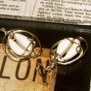 May include: A pair of silver earrings with a white cowrie shell encased in a wire circle. The earrings have a simple hook closure.