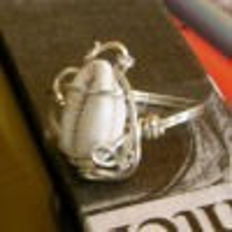 May include: A silver ring with a white oval-shaped stone set in a delicate, swirling design.