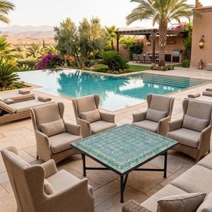 May include: A square outdoor table with a green mosaic tile top and black metal legs, surrounded by six beige upholstered wingback chairs on a stone patio next to a rectangular swimming pool with clear blue water and palm trees.