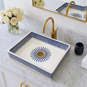 Puede incluir: Un lavabo rectangular de cerámica blanca con un borde a rayas azules y un diseño de rayos de sol alrededor del desagüe. El lavabo está sobre una encimera blanca con un grifo dorado y una taza negra.