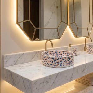 Peut inclure: Une vasque ronde en céramique ornée d'un motif peint à la main de feuilles délicates bleues et roses sur fond blanc, posée sur un comptoir en marbre blanc avec un robinet doré et un miroir octogonal assorti avec éclairage intégré.