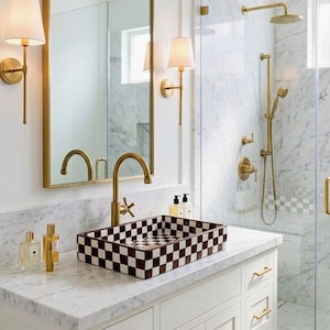 Op de afbeelding: Een moderne badkamer met een wit marmeren aanrechtblad en een rechthoekige wastafel met bruin-witte dambordpatroon. De kraan en wandkandelaars zijn goudkleurig. De douche heeft een glazen deur en gouden douchekranen.