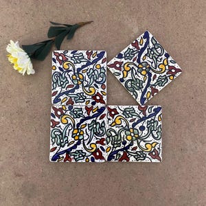 Puede incluir: Cuatro posavasos cuadrados de cerámica con un diseño floral en rojo, amarillo, verde y azul sobre un fondo blanco. Un posavasos tiene forma de diamante. Los posavasos están dispuestos sobre una superficie marrón con una flor y hojas decorativas.