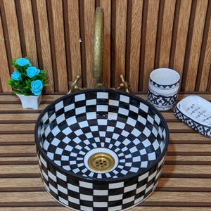 Peut inclure: Une vasque ronde en céramique avec un motif à damier noir et blanc à l'intérieur et à l'extérieur, dotée d'une bonde en laiton. Le lavabo est accompagné d'un petit pot blanc avec des fleurs bleues, d'un gobelet à motifs et d'un porte-savon.