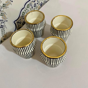 Peut inclure: Quatre tasses en céramique à rayures noires et blanches et bordures dorées. Les tasses sont disposées sur une surface blanche, avec une assiette décorative en arrière-plan. Les tasses ont une forme cylindrique.