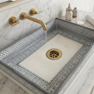 May include: A rectangular ceramic sink with a decorative grey and white geometric pattern. The sink has a brass drain and is mounted with brass fixtures. Water flows from the brass tap into the sink.