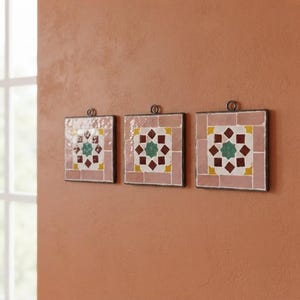 Peut inclure: Trois carreaux muraux carrés présentant un motif géométrique en étoile central dans des tons rouge foncé, vert et jaune, entouré d'une bordure de carreaux rectangulaires rose clair. Chaque carreau possède un petit anneau métallique au sommet.