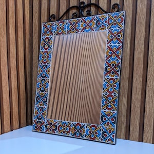 May include: A rectangular wall mirror featuring a decorative border of square tiles with a repeating floral pattern in red, blue, and yellow. The mirror has a black metal frame with a scrollwork top detail and a reflective surface showing vertical lines.