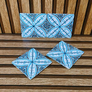 Op de afbeelding: Vier vierkante keramische onderzetters met een symmetrisch blauw en wit bloemmotief. Twee onderzetters staan rechtop naast elkaar, terwijl de andere twee plat in een ruitvorm zijn geplaatst, wat het gedetailleerde decoratieve ontwerp toont.