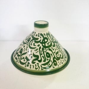 May include: A ceramic tagine pot with a conical lid and a circular base. The pot is off-white with green trim and features green Arabic calligraphy. The tagine is a traditional Moroccan cooking vessel.
