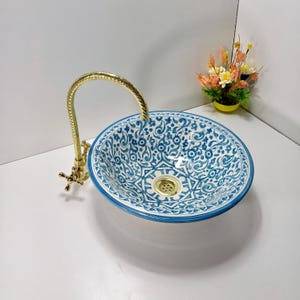 以下が含まれることがあります： 青と白の花柄模様の丸いセラミック洗面台。洗面台には、曲がった注ぎ口と十字型のハンドルが付いた金色の蛇口が付いています。洗面台は、カウンターの上に置くように設計されたベッセルシンクです。 洗面台の直径は約30cmです。