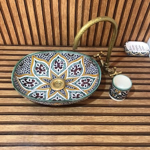 May include: An oval ceramic vessel sink featuring a hand-painted floral pattern in blue, yellow, and dark red. It includes a tall, curved brass tap with two handles, a matching cylindrical toothbrush holder, and a rectangular soap dish.