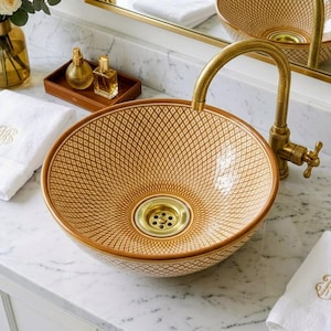 May include: A round, decorative bathroom sink with a gold and brown geometric pattern. The sink has a gold faucet and drain. White towels and perfume bottles are visible on the marble countertop.