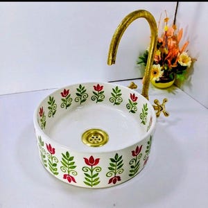Peut inclure: Un lavabo rond en céramique blanche orné d'un motif floral de tulipes rouges et de feuilles vertes. Il est équipé d'un robinet et d'un drain dorés.