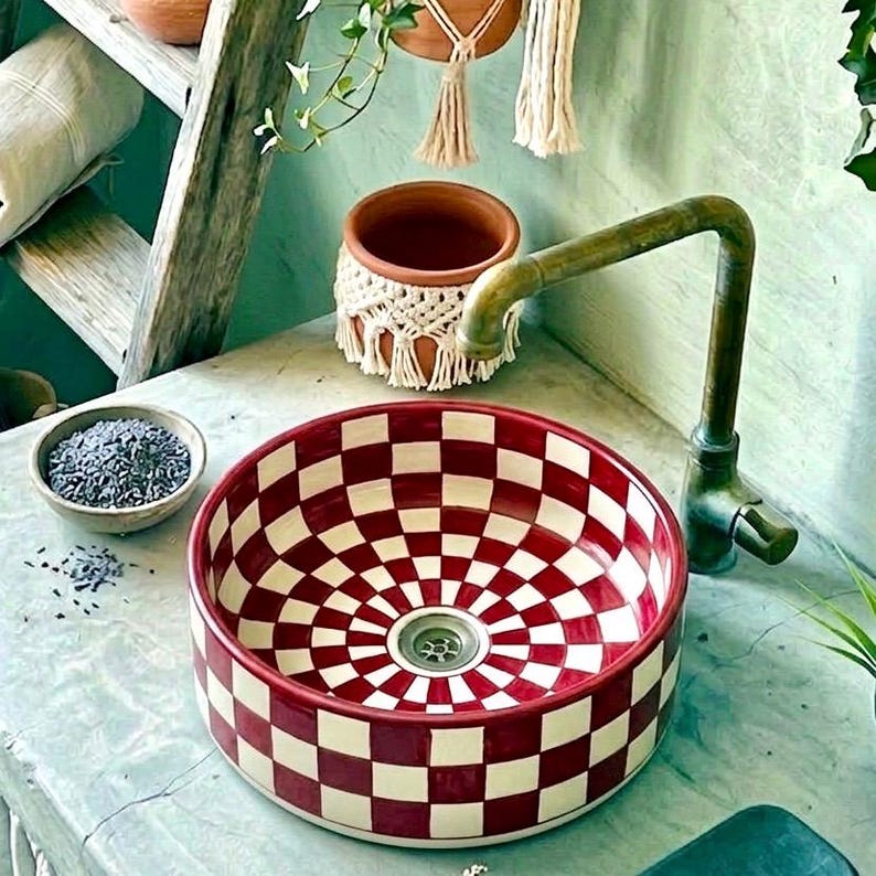 May include: A round ceramic sink with a red and white checkerboard pattern. The sink has a brass faucet and is surrounded by decorative elements, including a small bowl and a macrame pot.