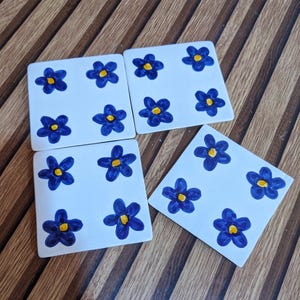 Peut inclure: Un ensemble de quatre dessous de verre carrés en céramique blanche, chacun orné d'un motif peint à la main de quatre fleurs bleu foncé à cinq pétales avec des cœurs jaune vif. Les dessous de verre sont disposés en groupe.