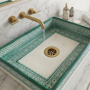 May include: Rectangular ceramic sink with a white interior and a green geometric border design. The sink has a brass drain and is mounted with brass fixtures, including a faucet and two handles. Water flows from the faucet into the sink.