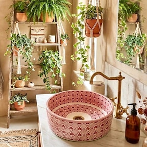 Puede incluir: Un baño con un lavabo rosa estampado y un grifo dorado. Plantas en macetas cuelgan del techo y se colocan en una estantería de escalera de madera. La habitación tiene un estilo natural y bohemio, con plantas y tonos neutros.