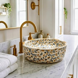 May include: A round, cream-colored ceramic bathroom sink with a floral pattern in blue and gold. The sink sits on a marble countertop next to a gold faucet and soap dispenser. White towels are stacked on the counter.