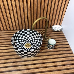 Peut inclure: Une vasque ronde en céramique avec un motif à damier noir et blanc, accompagnée d'un robinet haut et courbé en laiton antique. Un petit porte-savon assorti et un gobelet cylindrique au motif graphique coloré sont placés à proximité.