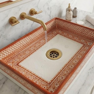 Puede incluir: Lavabo rectangular de cerámica con un patrón geométrico decorativo en tonos marrones y blancos. El lavabo tiene un desagüe de latón y está montado con un grifo de latón. El agua fluye del grifo al lavabo.