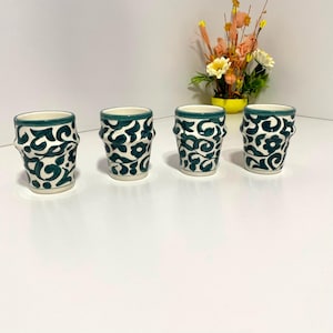Peut inclure: Quatre verres à shot en céramique avec une base blanche et un motif floral turquoise. Les bords sont turquoise et les verres ont un design texturé et incurvé. Un petit bouquet de fleurs est en arrière-plan.