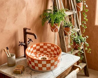 May include: A bathroom sink with a red and white checkered pattern. The sink is on a marble countertop with a bronze faucet. A wooden ladder holds hanging potted plants. A toothbrush holder and soap are also visible.