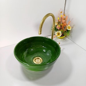 Puede incluir: Un lavabo de cerámica verde con un grifo dorado. El lavabo tiene forma redonda y un acabado brillante. El grifo tiene un diseño arqueado. Un pequeño ramo de flores está cerca.