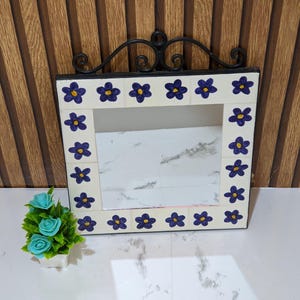 Puede incluir: Un espejo de pared cuadrado con un marco de azulejos blancos decorado con flores moradas pintadas a mano con centros amarillos. La parte superior del espejo está acentuada con un detalle de hierro forjado negro. El espejo tiene un centro de cristal.