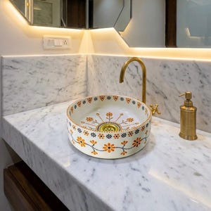 Peut inclure: Une vasque ronde en céramique à fond blanc ornée de motifs floraux orange et noirs, accompagnée d'un robinet haut en laiton et d'un distributeur de savon assorti, posée sur un plan de travail en marbre blanc aux veines grises.