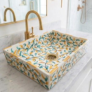 Op de afbeelding: Rechthoekige keramische badkamer wastafel met een bloemenpatroon in teal, geel en goud. De wastafel heeft een gouden afvoer en rand, en staat op een wit aanrechtblad. Een gouden kraan is zichtbaar op de achtergrond.