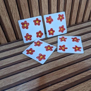 Peut inclure: Un ensemble de quatre dessous de verre carrés en céramique blanche, chacun orné d'un motif peint à la main de quatre fleurs rouges aux centres jaunes. Les dessous de verre ont une finition brillante et sont disposés en groupe.