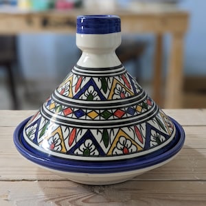 Peut inclure: Un tajine en céramique avec un couvercle conique et une base large et peu profonde. Le pot est blanc avec un bord et un dessus de couvercle bleus. Le couvercle est décoré d'un motif géométrique bleu, rouge, jaune et vert.