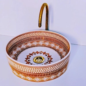 Puede incluir: Un lavabo redondo de cerámica blanca con intrincados patrones geométricos marrones. El lavabo tiene un grifo y un desagüe dorados. El diseño presenta un borde detallado y un medallón central, creando una estética elegante.