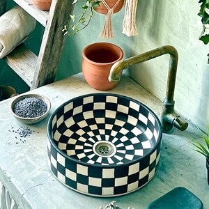 May include: A black and white checkered ceramic sink with a brass faucet. The sink has a circular design with a drain in the center. A small bowl of lavender sits nearby, along with a terracotta pot and hanging macrame.