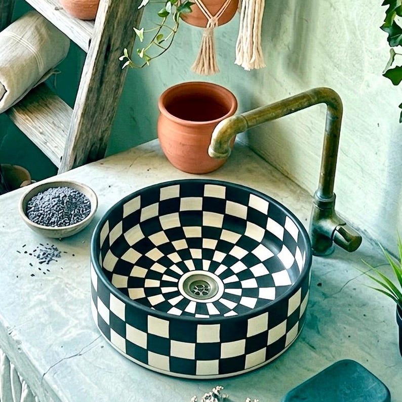 May include: A black and white checkered ceramic sink with a brass faucet. The sink has a circular design with a drain in the center. A small bowl of lavender sits nearby, along with a terracotta pot and hanging macrame.