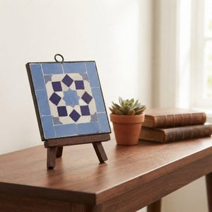 Peut inclure: Un carreau de mosaïque carré présentant un motif géométrique de carreaux bleu clair, blancs et bleu foncé disposés en étoile centrale. Le carreau est encadré d'un bord en métal sombre avec une petite boucle de suspension sur un chevalet en bois.