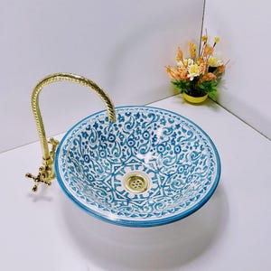 Peut inclure: Un lavabo rond en céramique avec un motif floral bleu et blanc. Le lavabo est équipé d'un robinet et d'un drain dorés. Un petit bouquet de fleurs dans un pot jaune est en arrière-plan. C'est un élément de salle de bain décoratif.