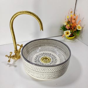 May include: A patterned ceramic sink in grey and white, with a gold-coloured drain and a matching gold tap. The sink has a decorative geometric design. A small floral arrangement is in the background.