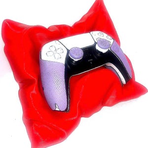 May include: A purple and white PlayStation 5 controller with purple buttons, resting on a red pillow.