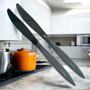 Puede incluir: Dos cuchillos de mantequilla de color gris oscuro con mangos texturizados se apoyan en un armario blanco. Los cuchillos están angulados hacia el espectador, mostrando su elegante diseño. Una olla naranja y una olla de acero inoxidable son visibles en el fondo.