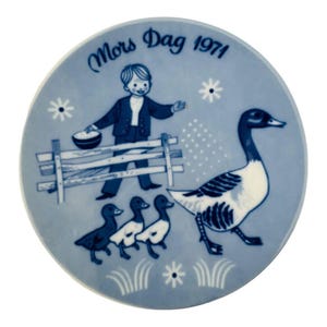 May include: A blue and white decorative plate with a pastoral scene. A boy in a blue suit stands near a fence, feeding a goose and three ducklings. The plate is inscribed with "Mors Dag 1971".